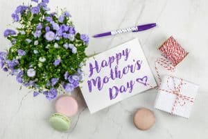 Flat lay of Mother's Day card with purple flowers, gift, macarons, and a pen indoors.