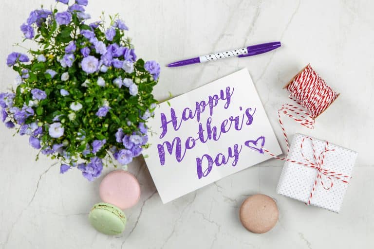 Flat lay of Mother's Day card with purple flowers, gift, macarons, and a pen indoors.
