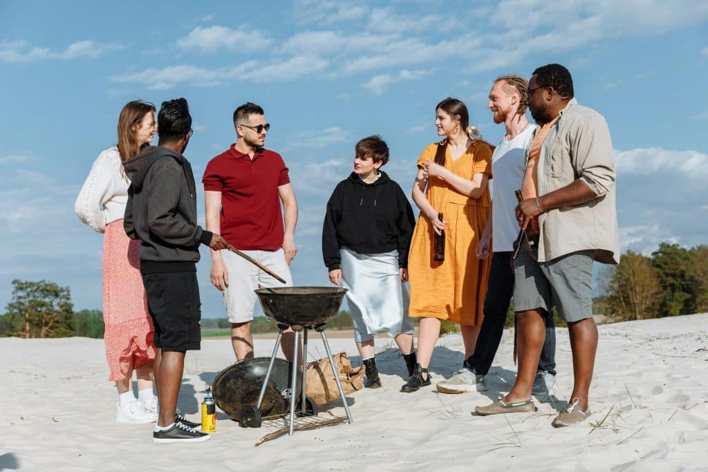 A diverse group of friends grilling together at the beach under a clear sky.