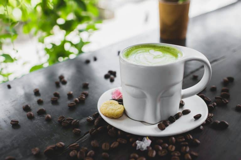 A vibrant matcha latte served with cookies on a saucer surrounded by coffee beans on a dark wooden surface.