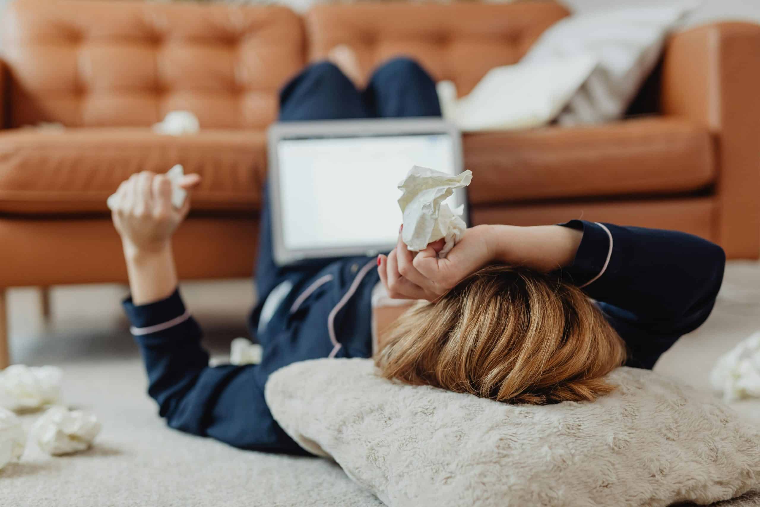 seasonal allergies, A woman in pajamas lies on the floor next to a laptop, surrounded by tissues, depicting a sick day at home.