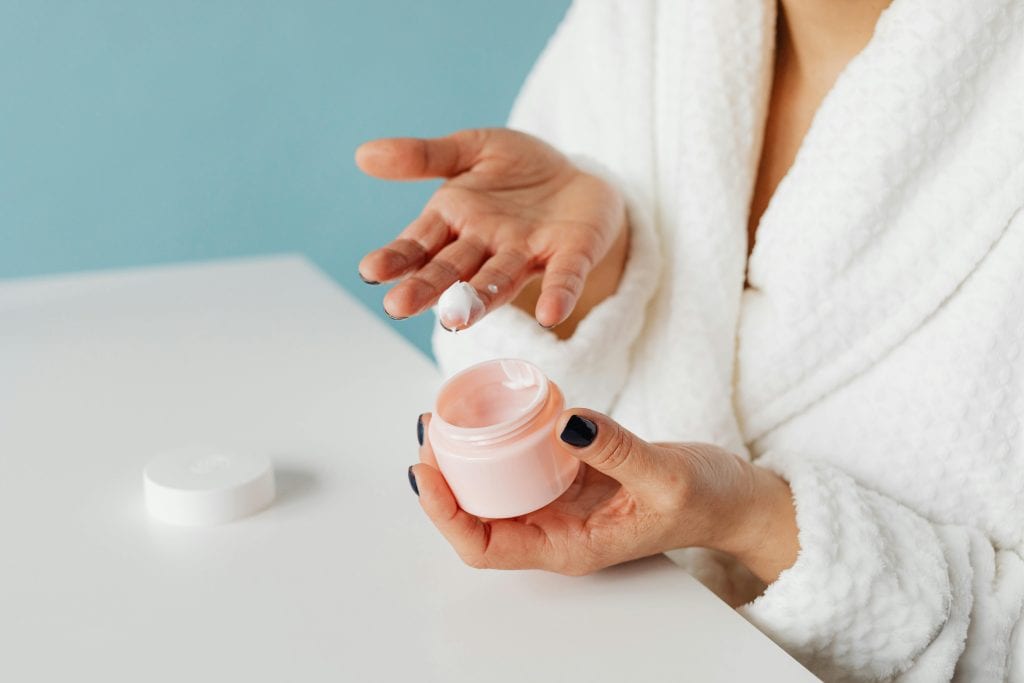 A woman in robe applying skincare cream from an open jar, highlighting moisturizing routine. body lotion