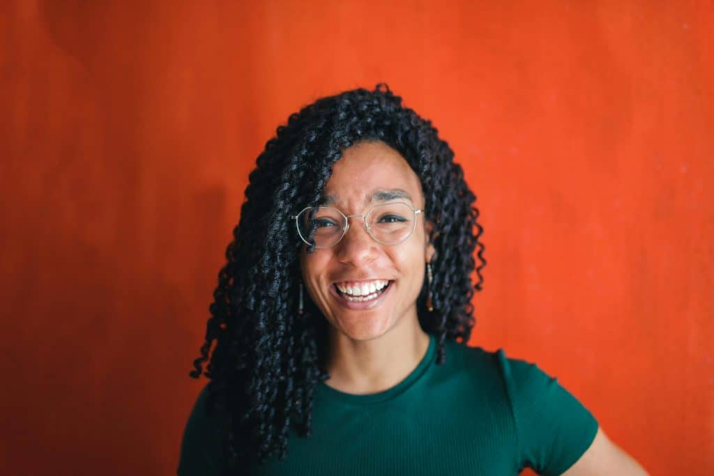 A joyful young woman with curly hair and glasses, smiling brightly against a vibrant red backdrop, daily devotion
