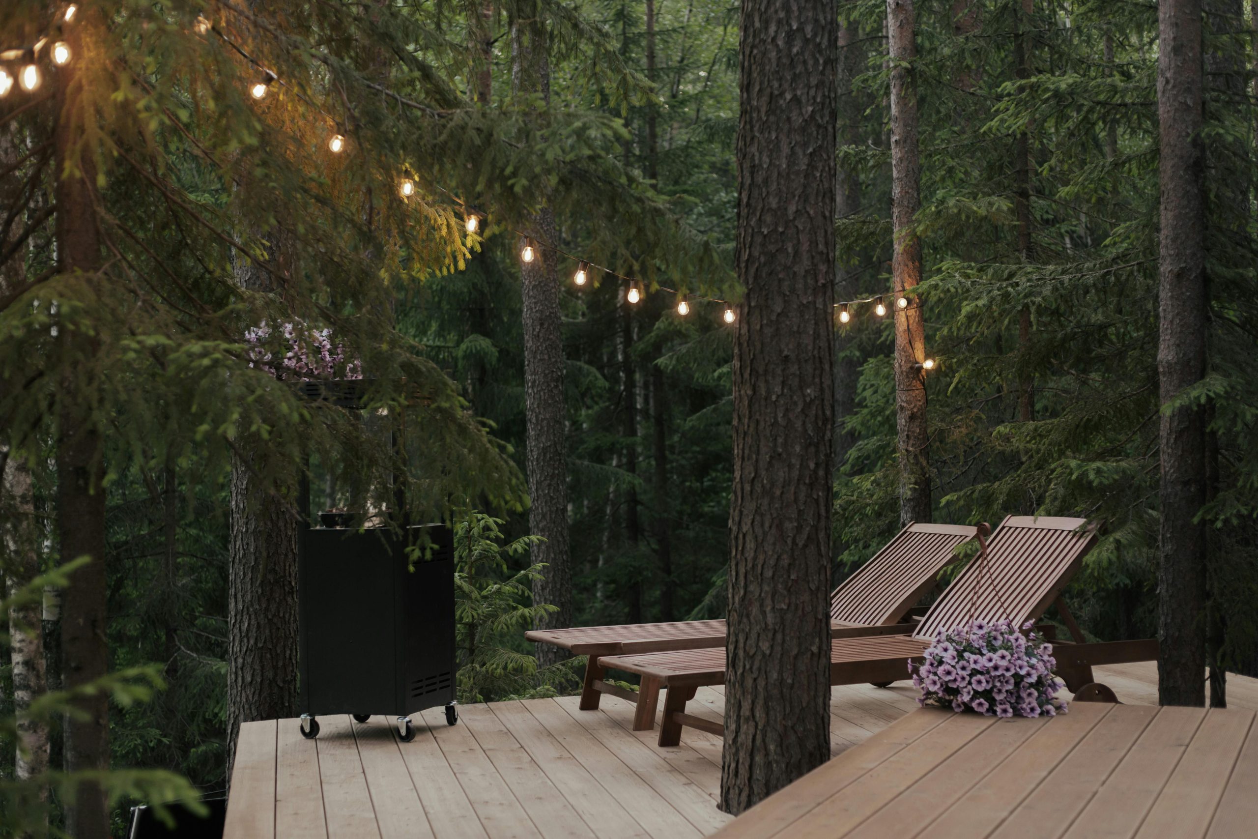 A peaceful forest scene featuring a wooden deck, sun loungers, and string lights.
