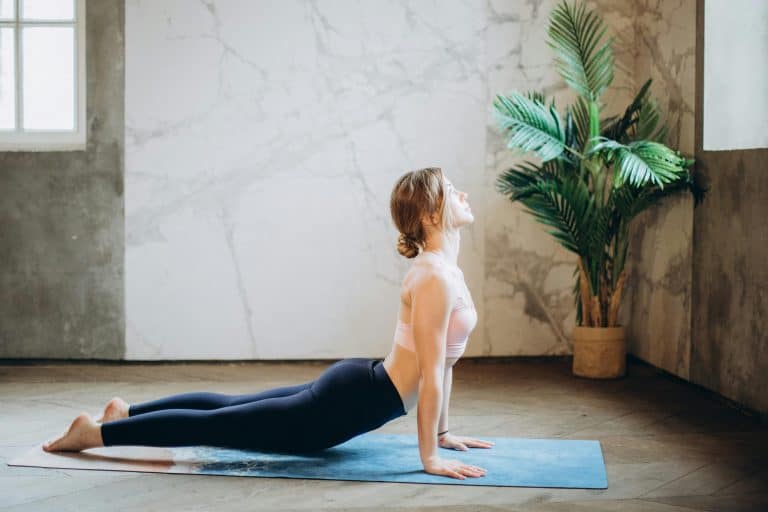 Woman in yoga pose on mat, exercising indoors with natural light and plant decor, somatic psychology, healing