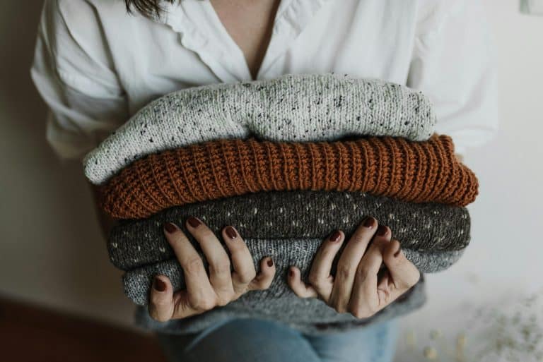A person holding a stack of folded knitted sweaters in various colors, with a focus on textiles and textures. Fall fashion, fashion trend