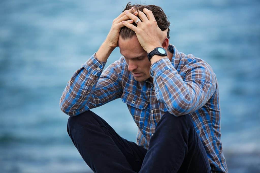 A man in a plaid shirt sits by the water looking distressed, symbolizing stress, daily devotion